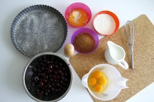 Plech, čerešne, rozbité vajcia v nádobe, mlieko, múka a ostatné potreby na pečenie koláča
